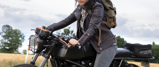 Alaqua Cox is seated on a black Royal Enfield Continental GT 650 motorcycle, parked outdoors with fields in the background and is looking straight into the camera. She is wearing a brown leather jacket, a plaid shirt, dark pants, and has a backpack on.